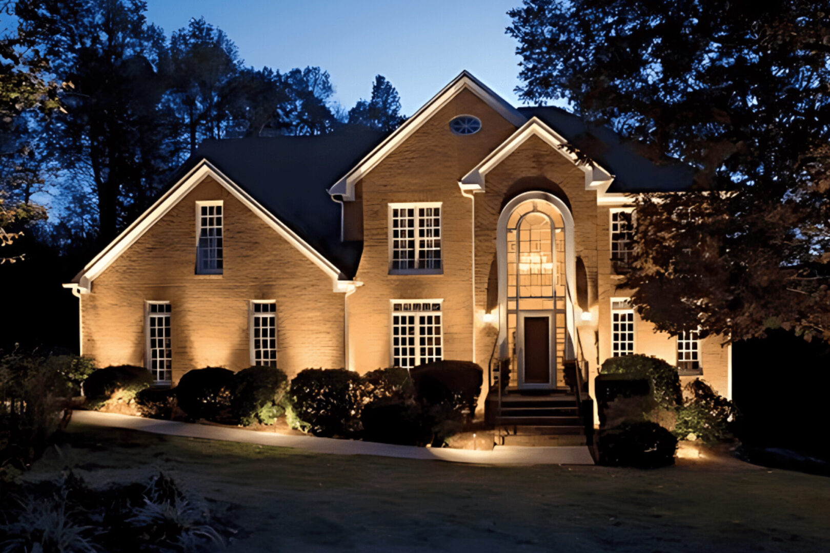 Two-story brick house with illuminated windows, curved front steps, and surrounding shrubs at dusk.
