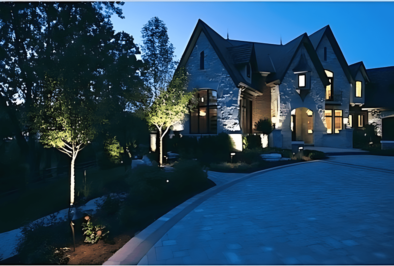 An illuminated large house with pointed roofs and stone exterior at dusk, surrounded by trees and a paved driveway.