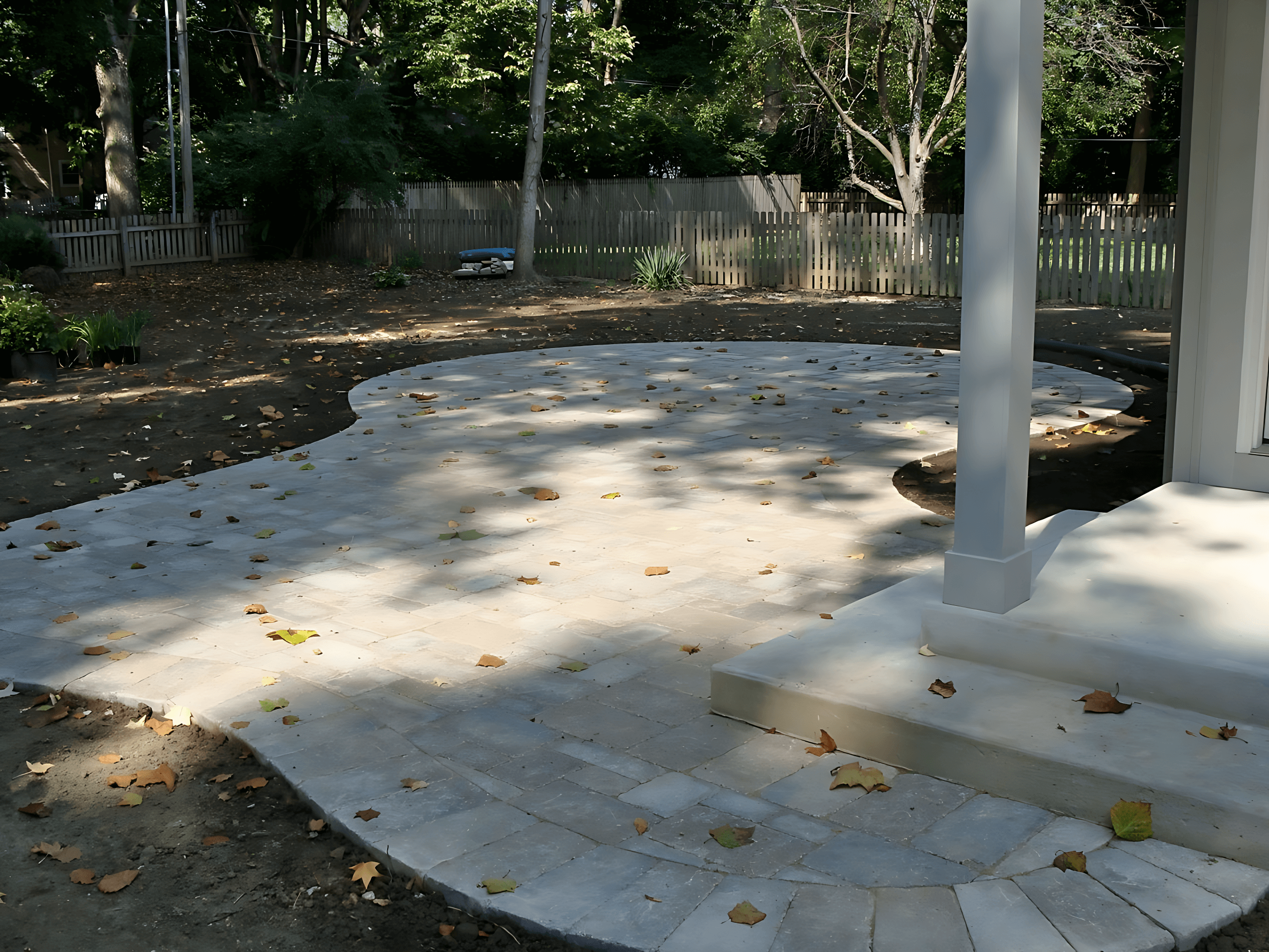 A newly constructed stone patio with fallen leaves, surrounded by trees and a wooden fence.