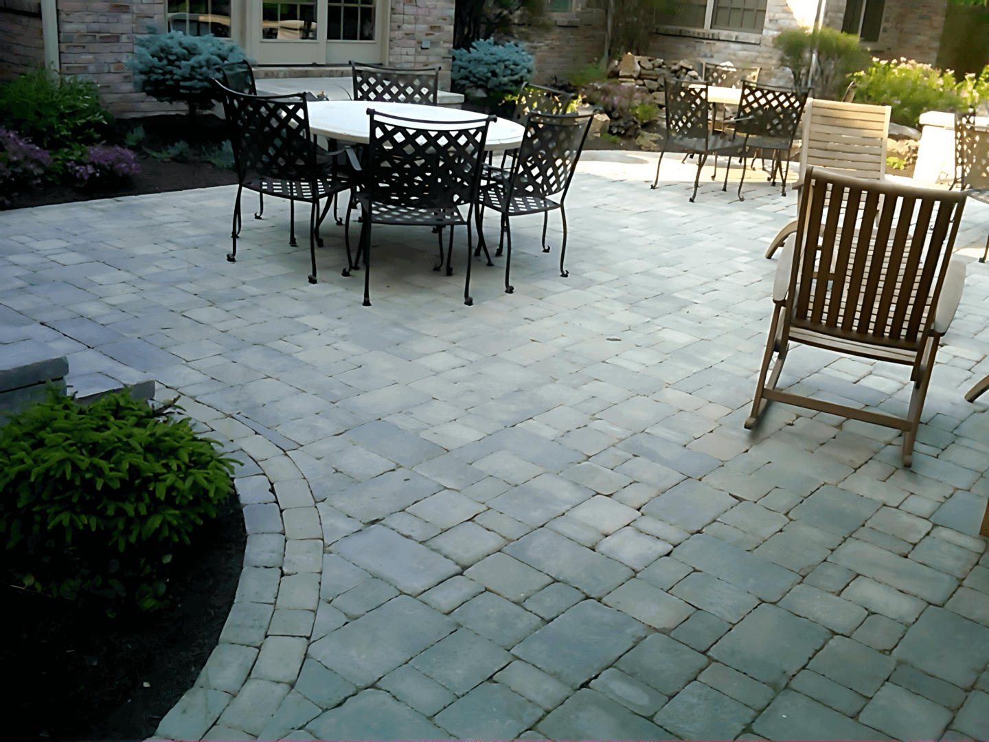 A patio with a table and six chairs, featuring a stone tile floor. A wooden chair is in the foreground, and there are plants around the perimeter.