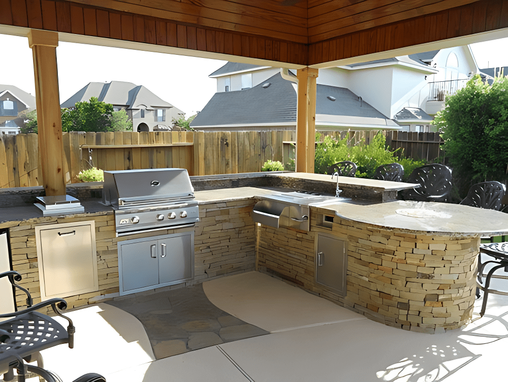Outdoor kitchen with stainless steel appliances, stone countertops, barbecue grill, and sink under a wooden pergola, surrounded by patio chairs and a wooden fence.