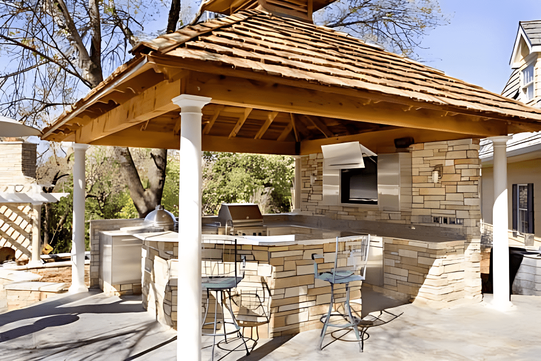 Outdoor kitchen with stone countertops, grill, and bar stools under a wooden pergola.