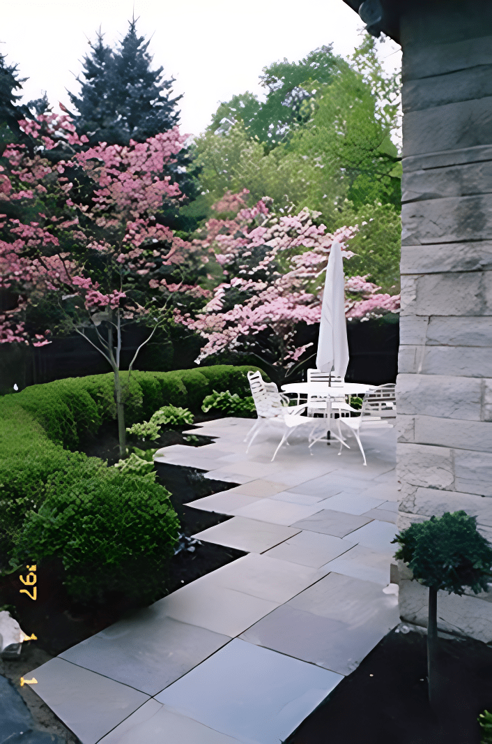 A stone patio with white furniture and an umbrella. Surrounded by neatly trimmed bushes and blooming pink trees. A stone building is partially visible on the right.