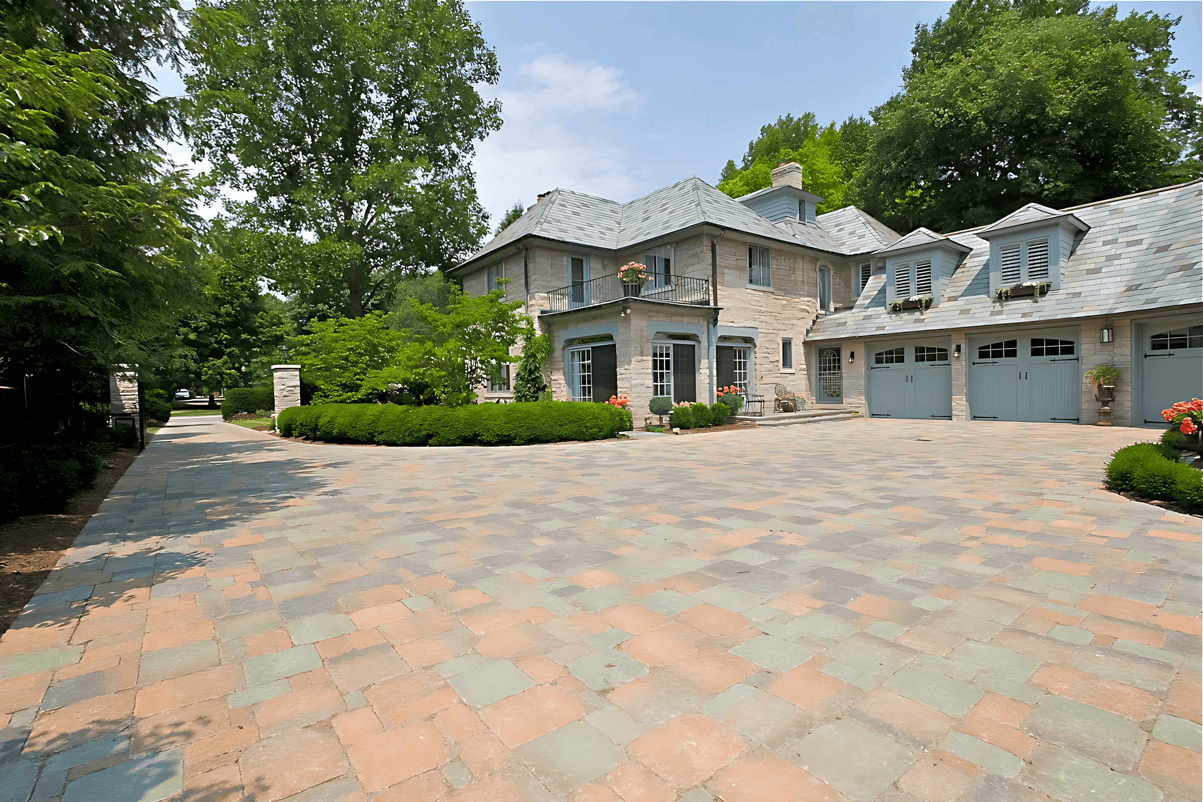 Large stone house with blue shutters and a three-car garage, surrounded by trees. The driveway is paved with multi-colored stones.