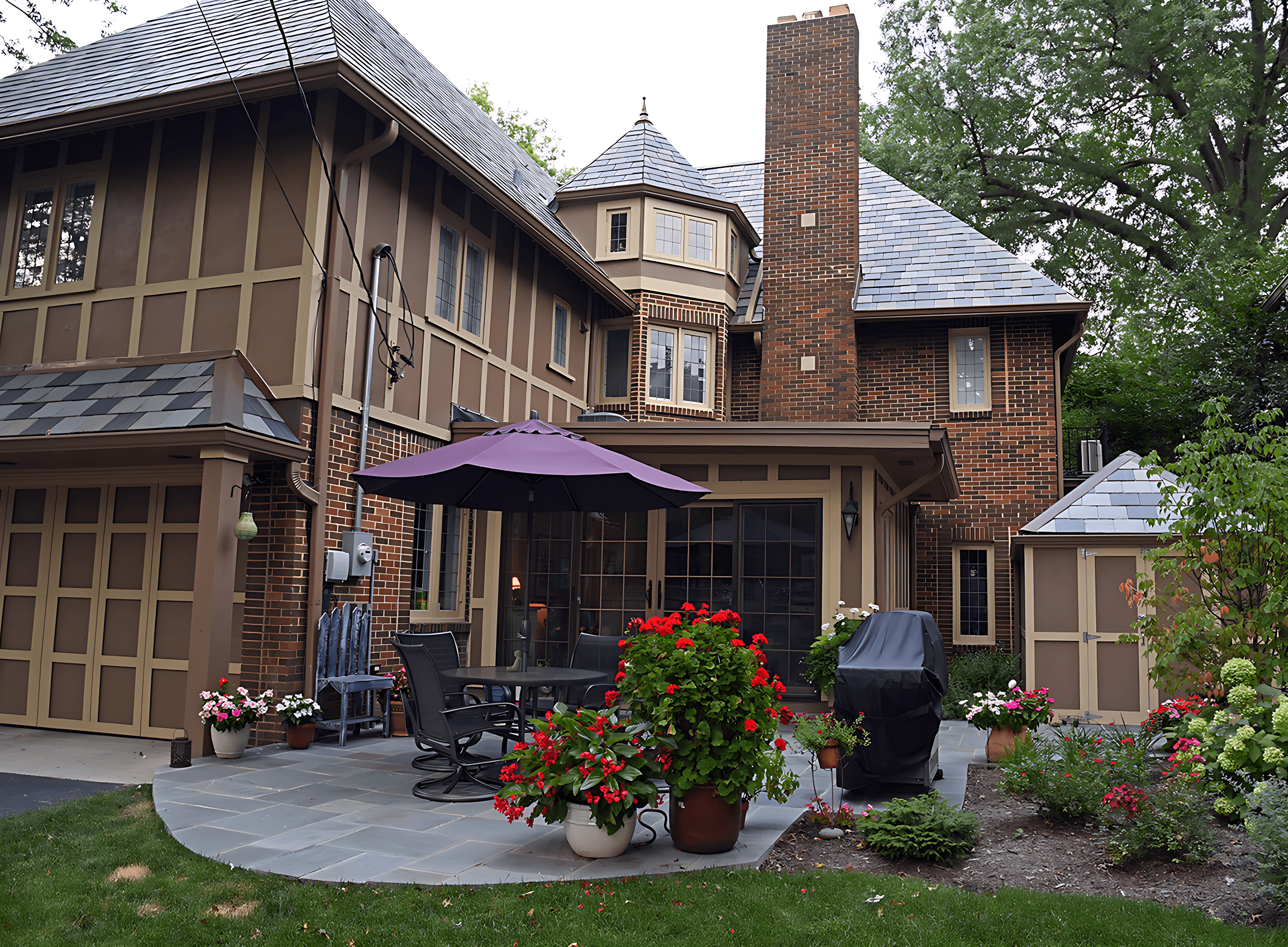 A brick house with a patio featuring outdoor furniture, a grill, potted plants, and an umbrella. The house has multiple stories and a prominent chimney.