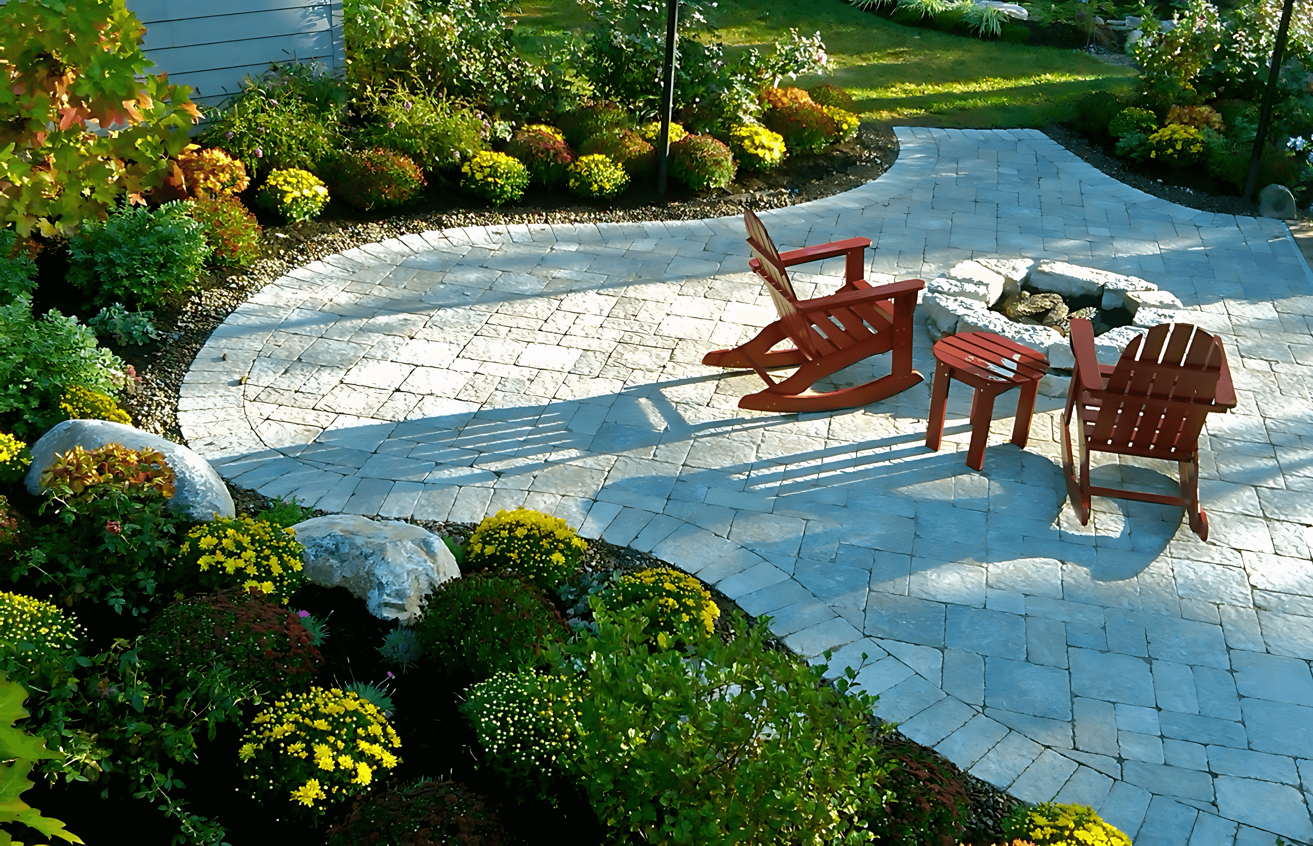 A garden with a stone patio features two wooden chairs and a small table. Colorful flowers and greenery surround the area.