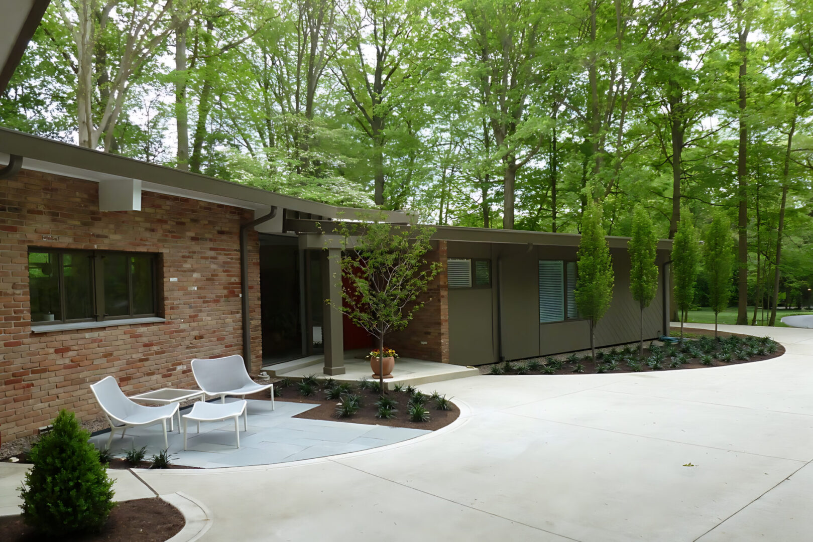 Modern house with brick and wood facade surrounded by trees. Two white chairs on a patio by the entrance, near a curved driveway.