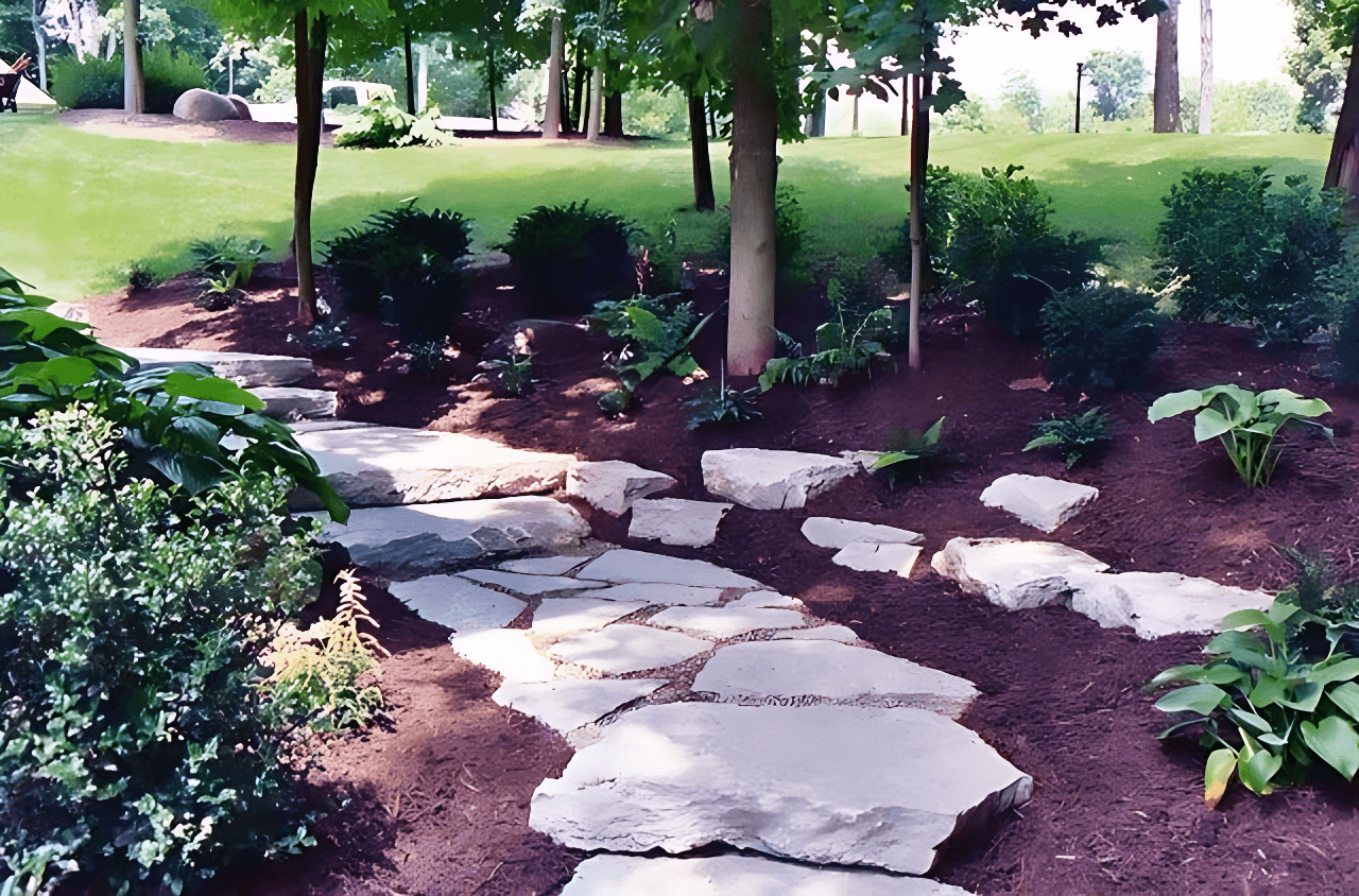 Stone path winding through a landscaped garden with trees, shrubs, and mulch under sunlight.