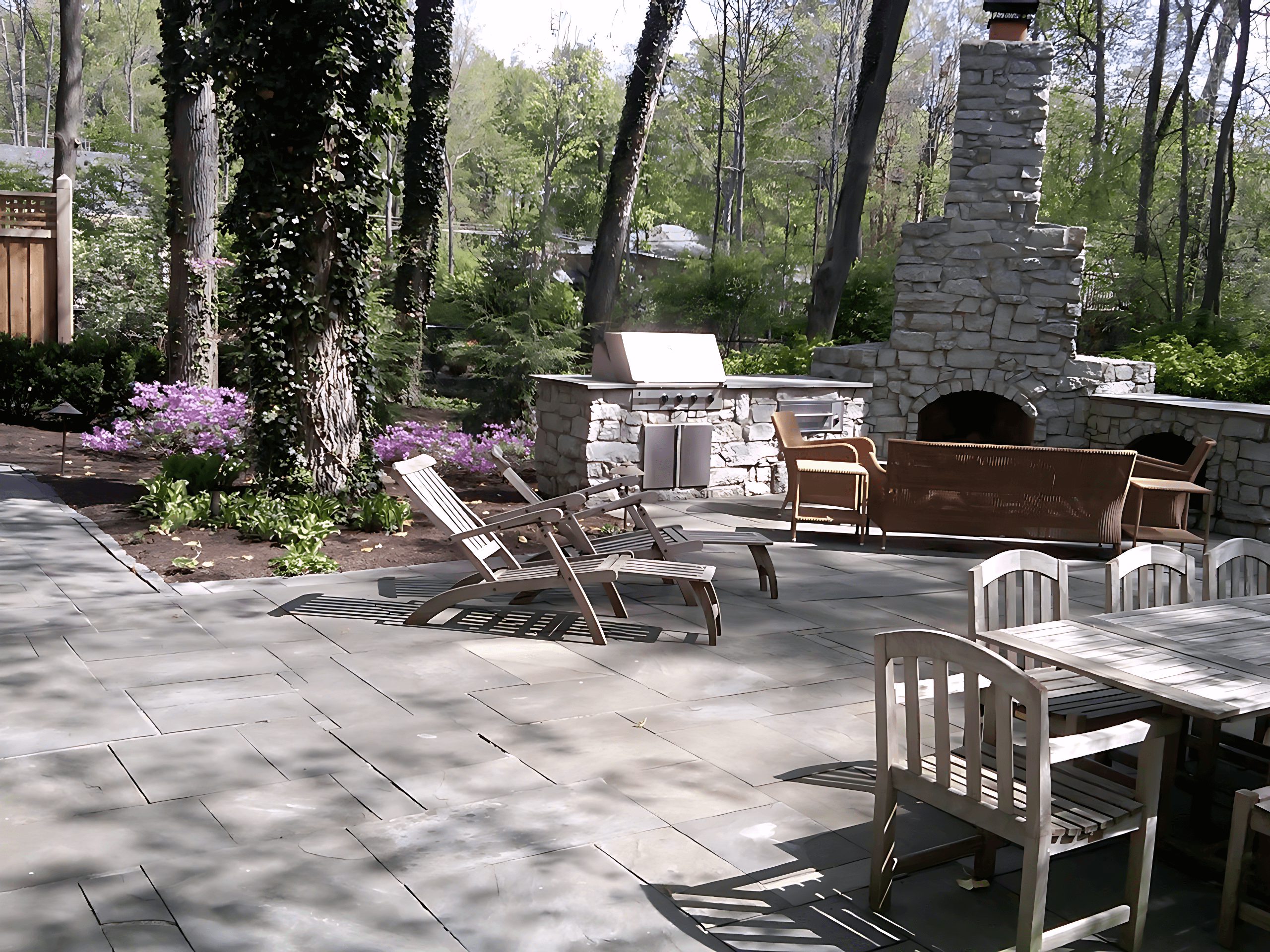 Stone patio with wooden chairs, dining table, outdoor fireplace, grill, and surrounding trees and flowers.