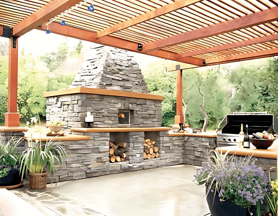 Outdoor stone fireplace under a wooden pergola, with stacked firewood, a grill, and surrounding plants.