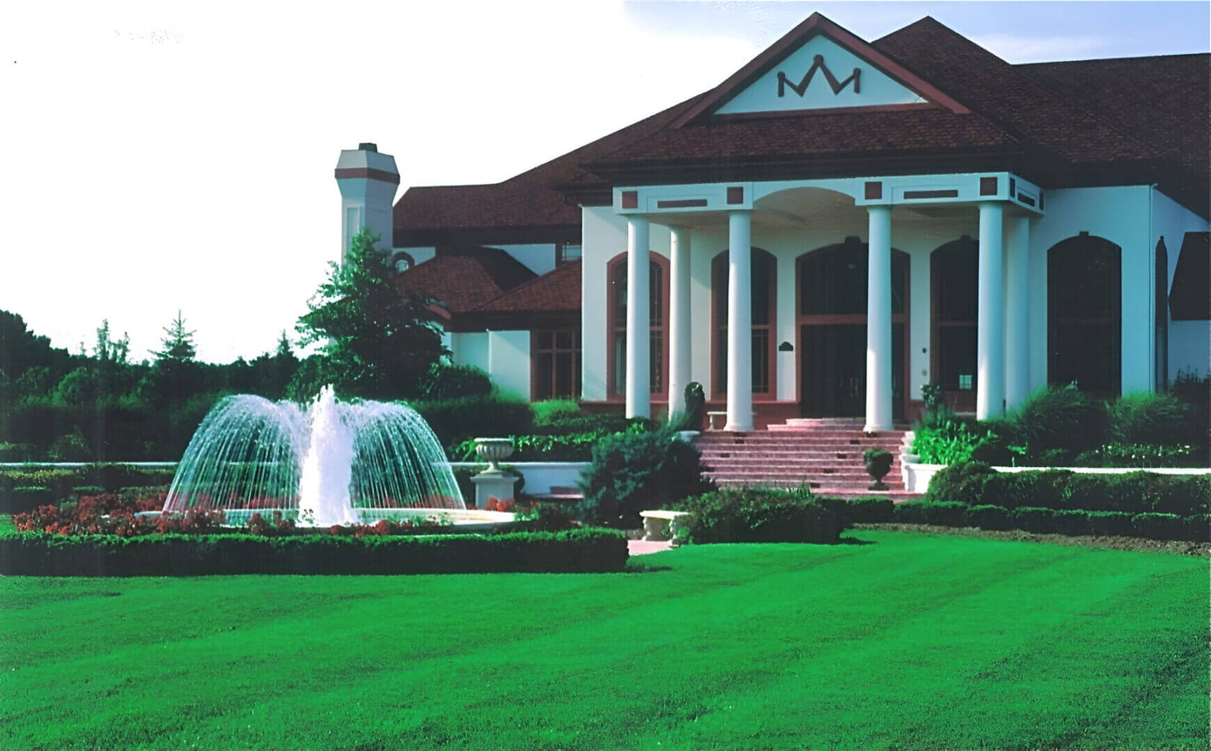 Large house with columns, red roof, and decorative "M" sign. Manicured lawn and garden with a fountain in front.