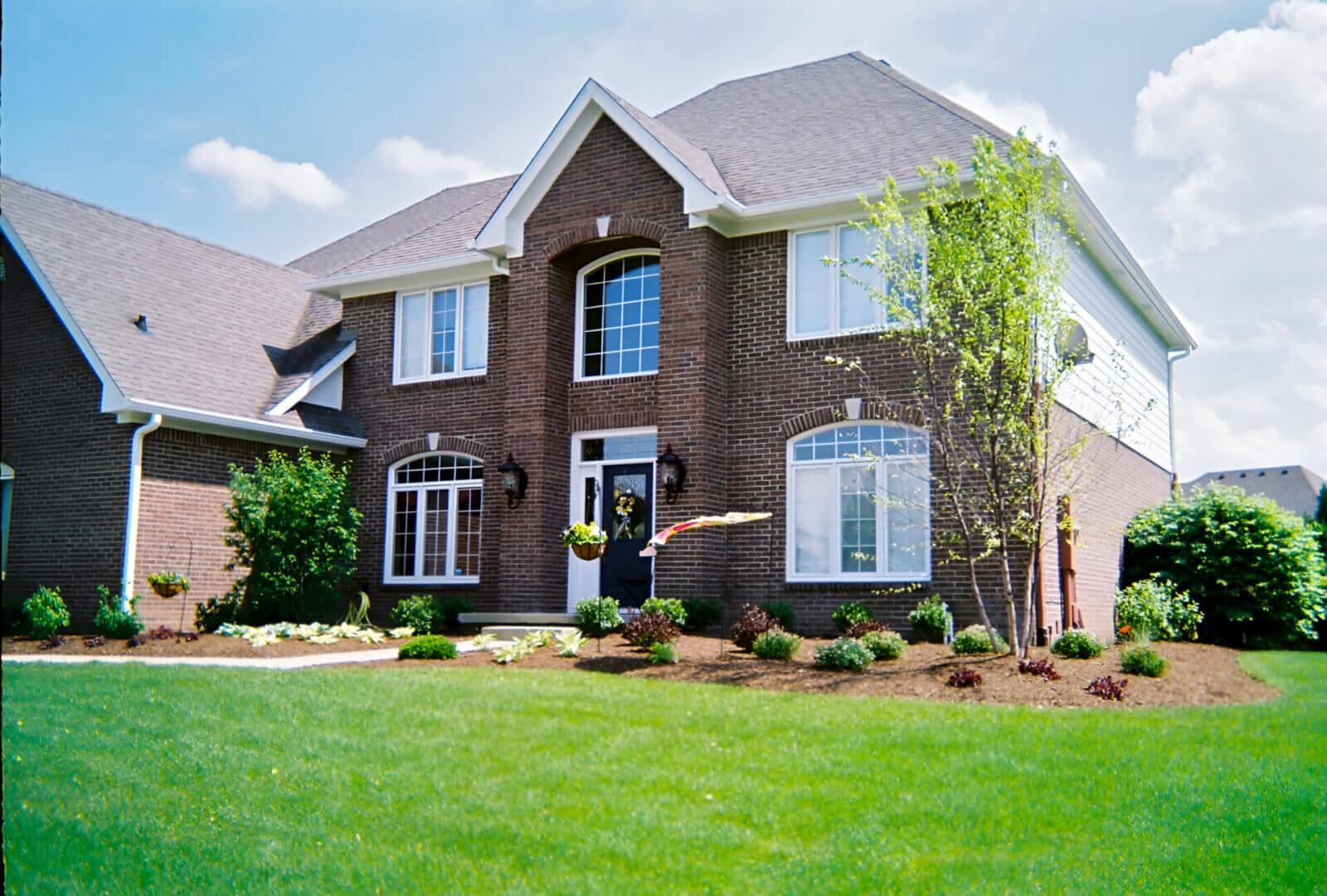 Two-story brick house with manicured lawn and small trees. Large arched window above the entrance and several smaller windows on the facade. Clear blue sky with few clouds in the background.