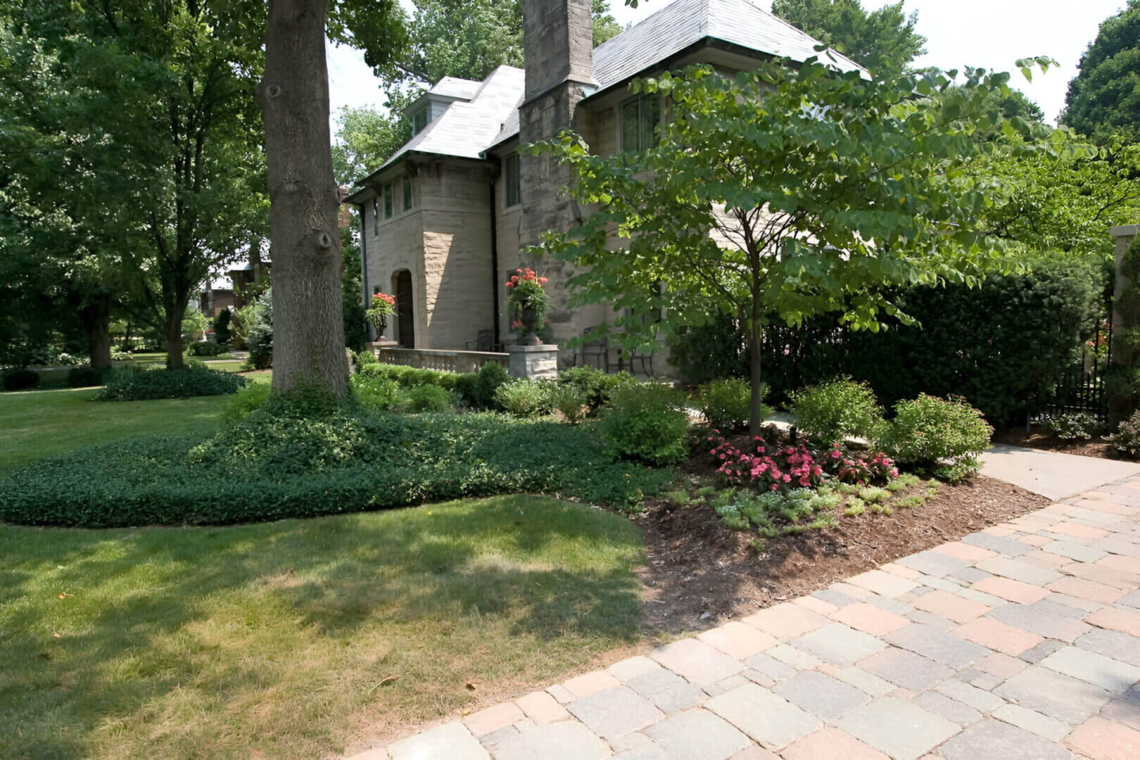 Stone house with a sloped roof surrounded by green trees and shrubs. A paved walkway and manicured lawn are in the foreground.