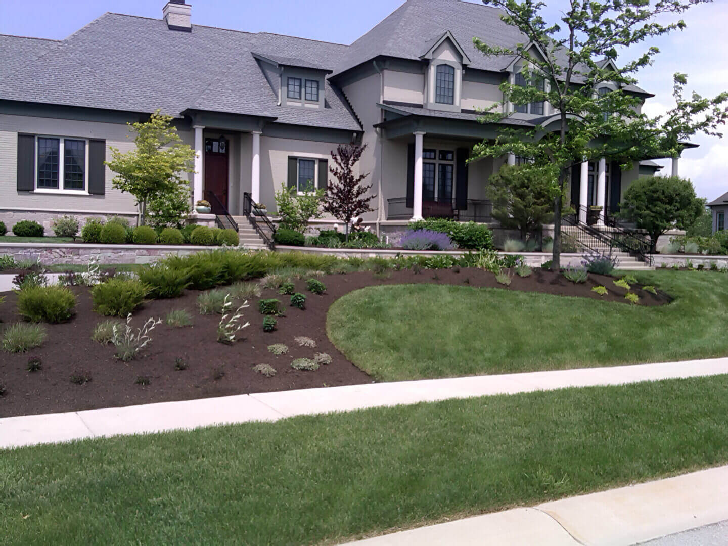 Large two-story house with gray siding and multiple gables. Manicured lawn and landscaped garden with shrubs and a small tree at the front. A sidewalk runs along the street.