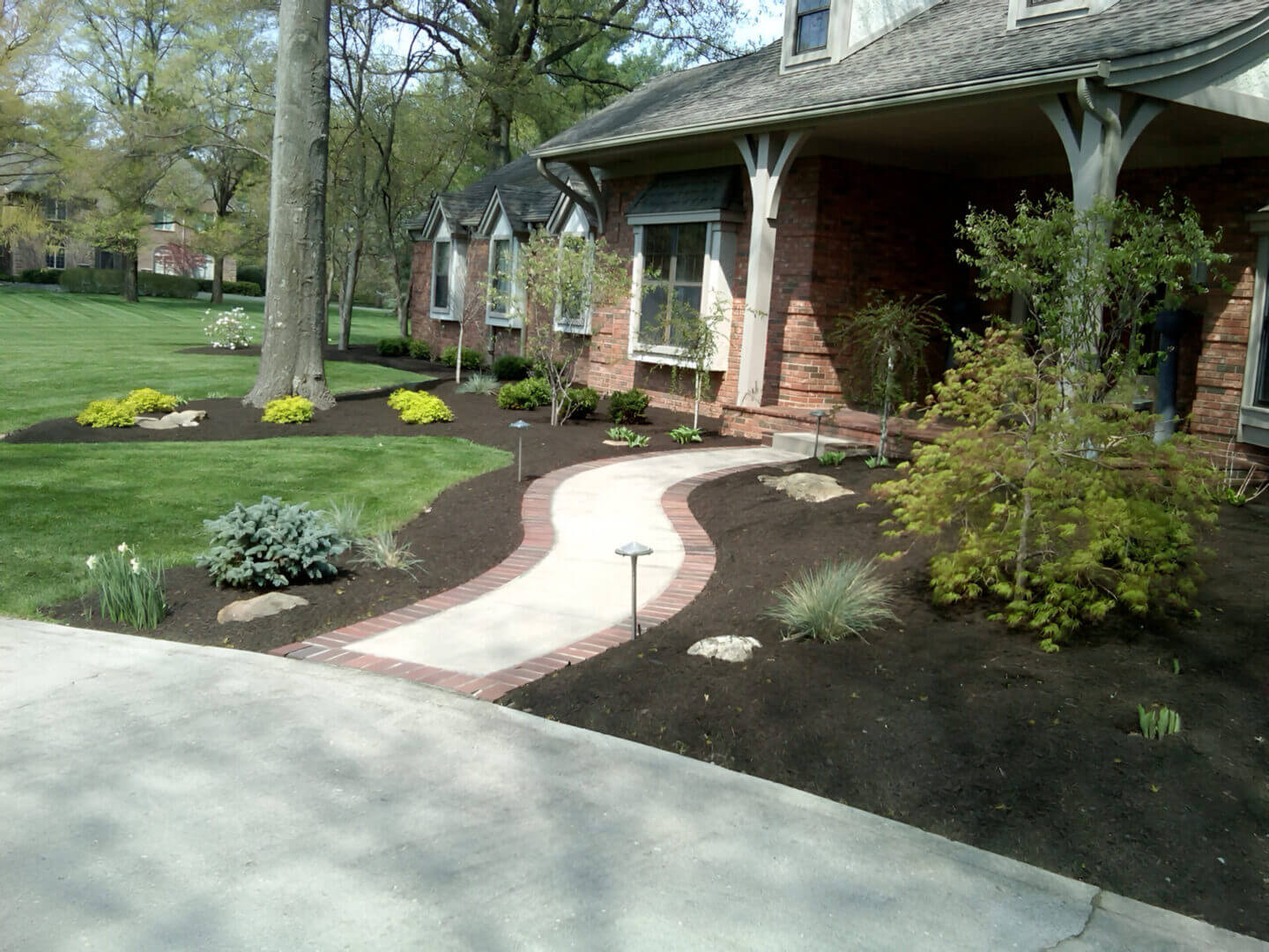A neatly landscaped front yard with a brick-edged path, mulch beds, small shrubs, and a manicured lawn in front of a brick house.