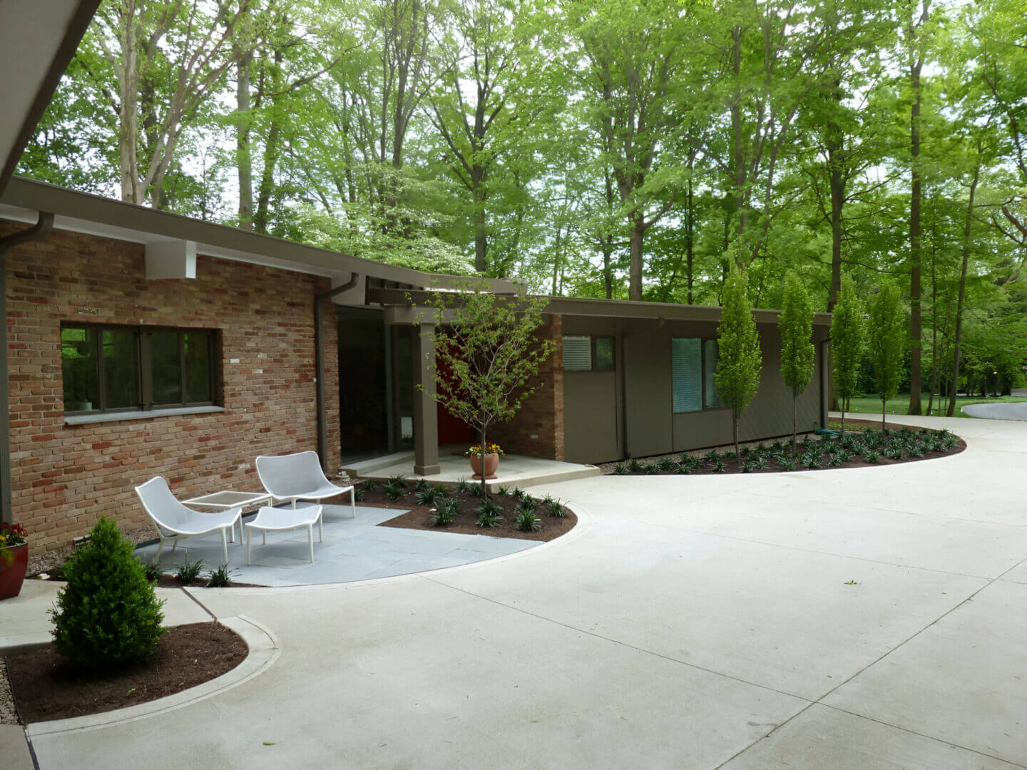 Modern house with brick and wood siding, surrounded by trees. Features a small patio with two white chairs and a round table on a concrete driveway.