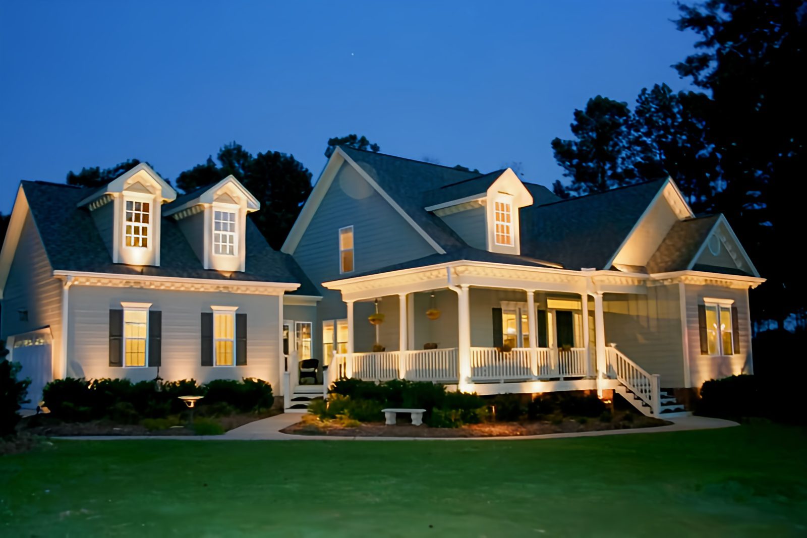 A well-lit, two-story house with a porch and gabled roof, surrounded by trees, at dusk.