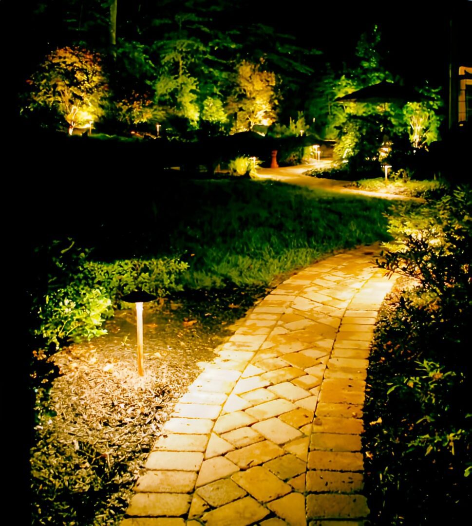 A brick pathway illuminated by ground lights curves through a garden at night, with surrounding trees and plants also lit up.