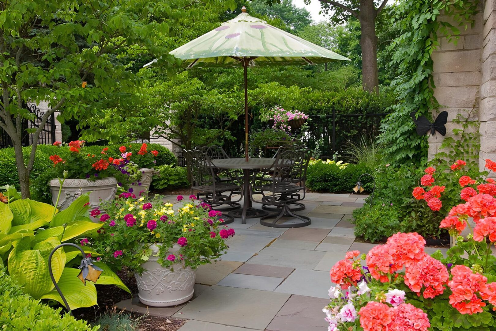 A garden patio with a metal table and chairs under a green umbrella, surrounded by blooming red and pink flowers and lush greenery.