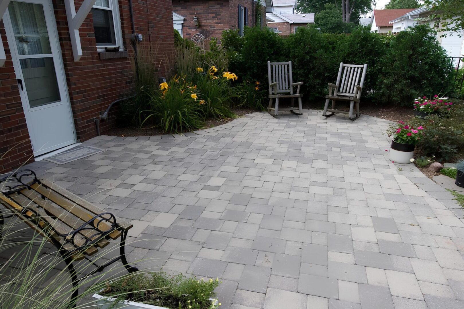 A brick patio with two wooden chairs, a bench, and potted plants. Yellow flowers and shrubs line the edge.