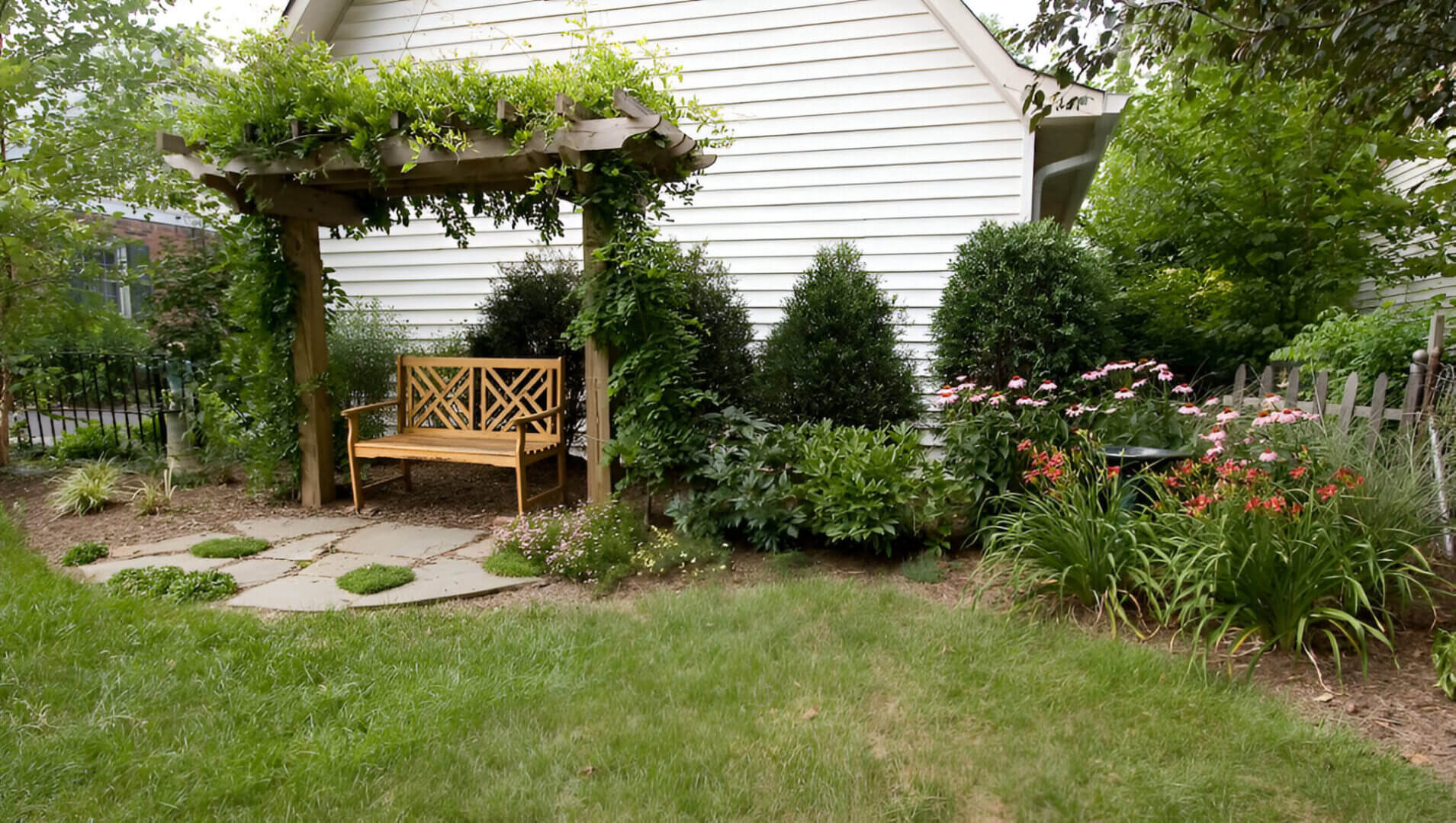 Wooden bench under a pergola with vines, surrounded by bushes and pink flowers, next to a white house wall in a garden setting.