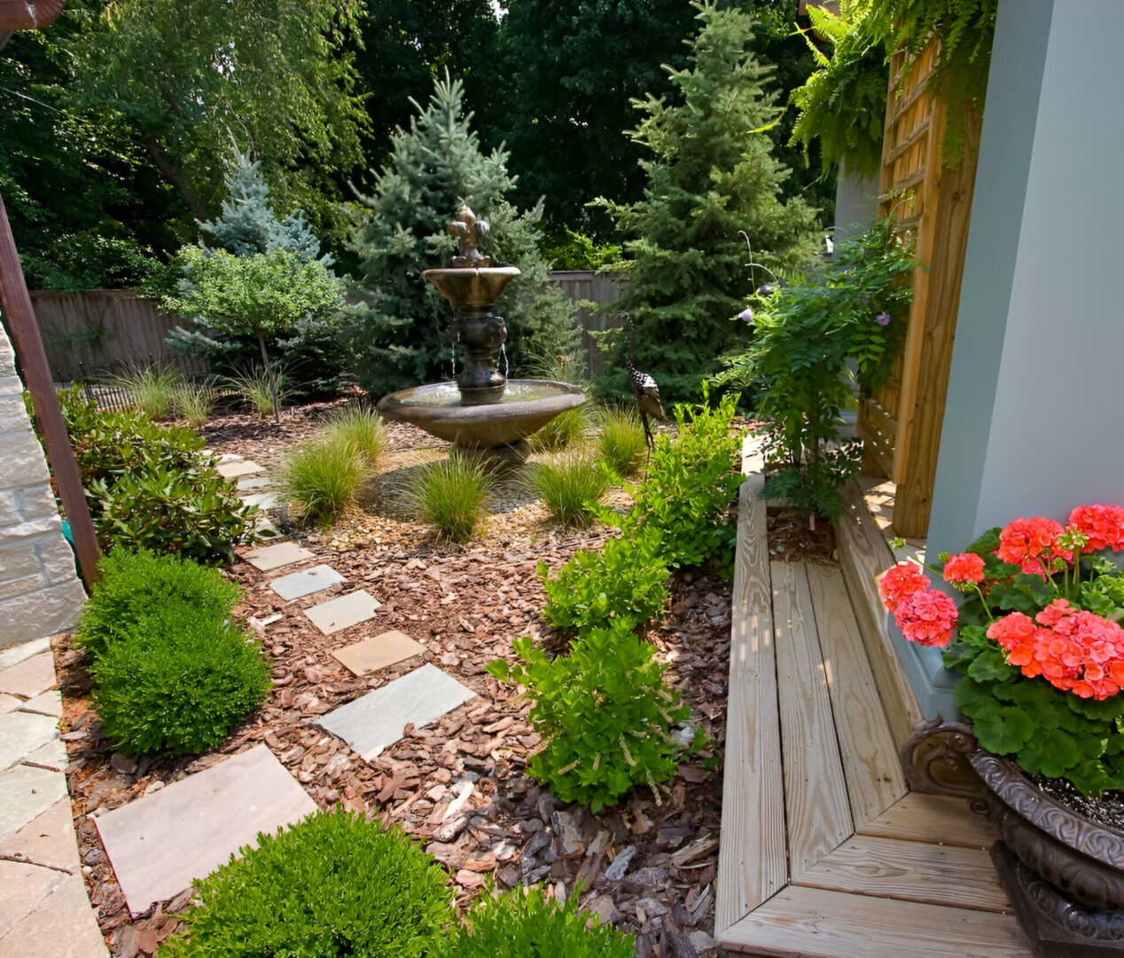 Garden with a stone path, central tiered fountain, various shrubs and flowers. Wooden bench with red geraniums on the right. Tall trees in the background.