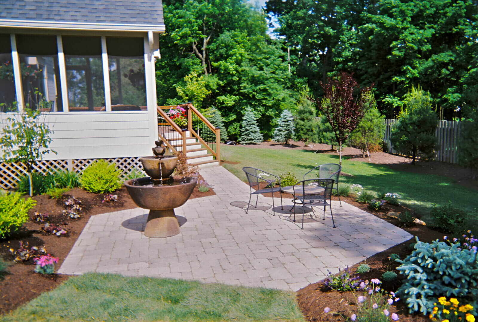 Backyard garden with a stone patio, a tiered fountain, two metal chairs, and a small table. Surrounded by grass, flowers, and trees.