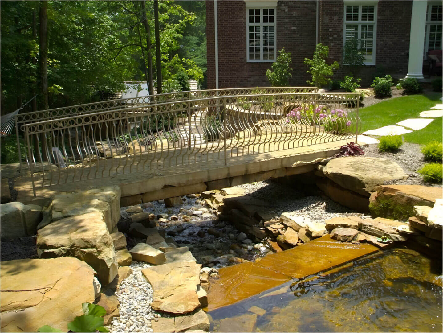 A small stone bridge with a decorative metal railing crosses over a rocky stream in a garden near a brick building.