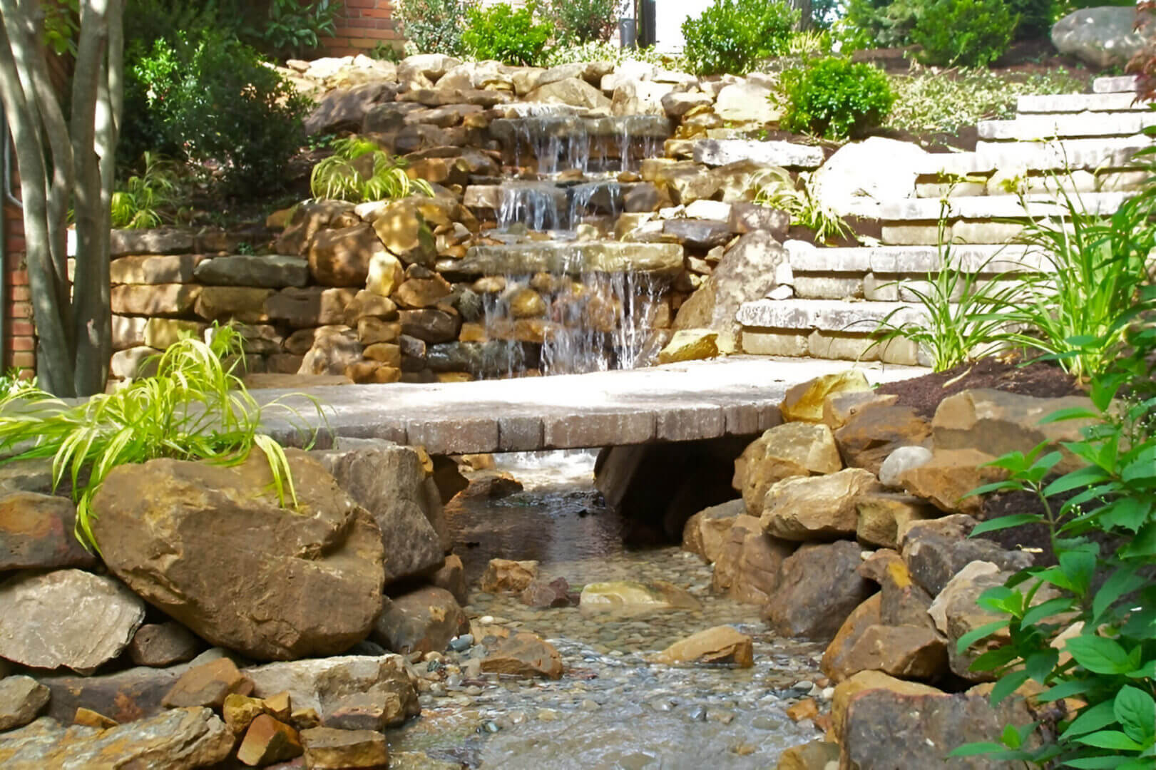 Stone waterfall feature with cascading water, rock steps, and a small bridge in a landscaped garden.