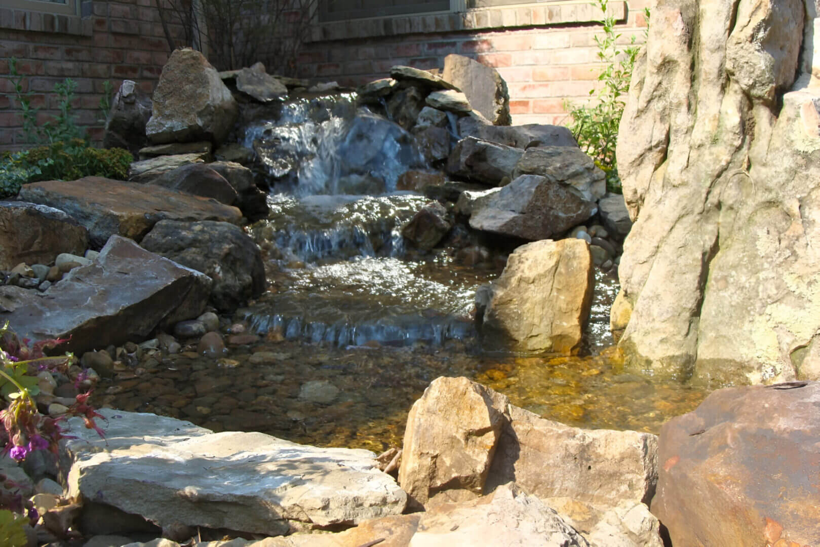 Small waterfall cascading over rocks into a clear pond, surrounded by stones, with a brick building in the background.
