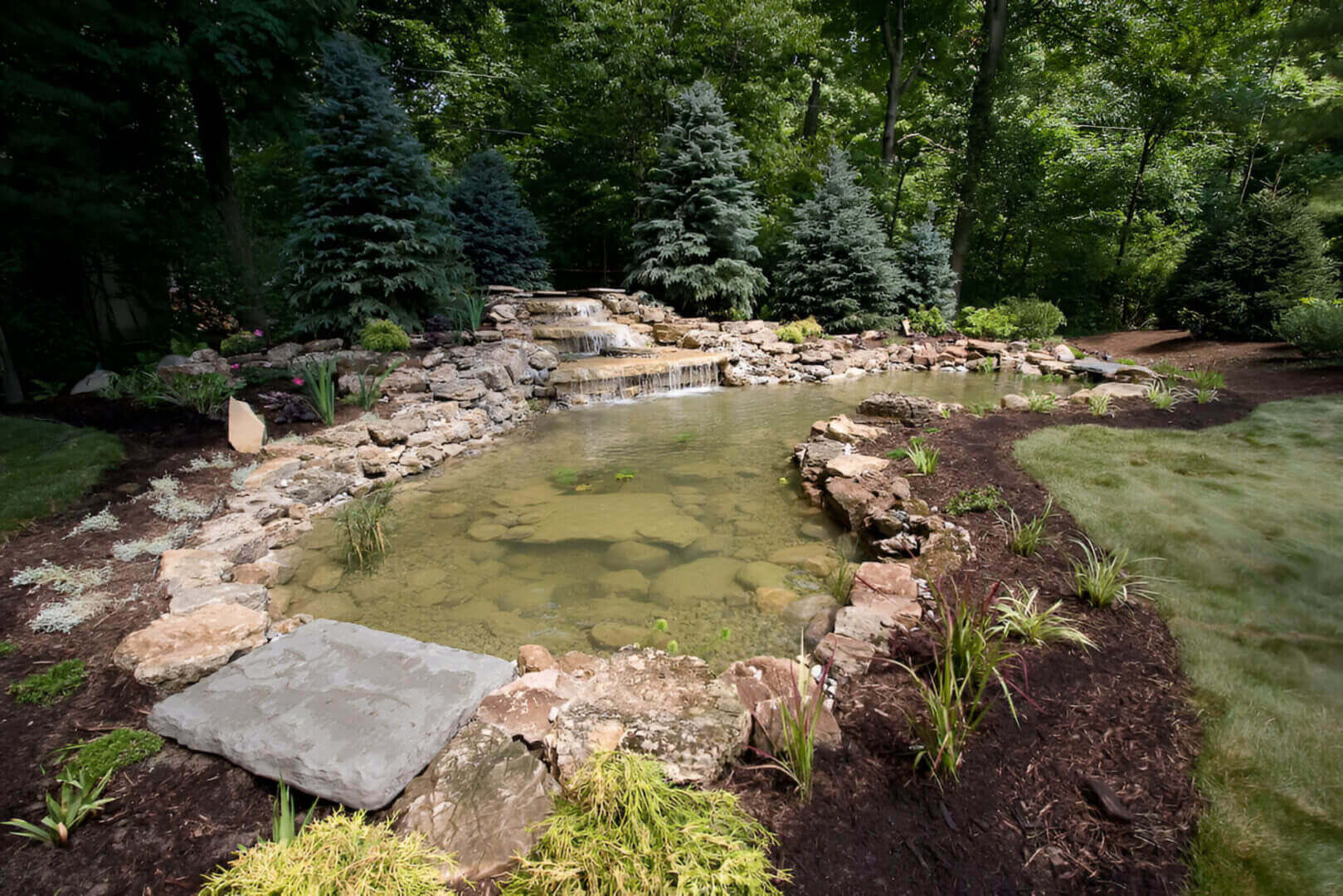 A landscaped garden features a pond with clear water, surrounded by rocks and greenery, with small waterfalls and pine trees in the background.