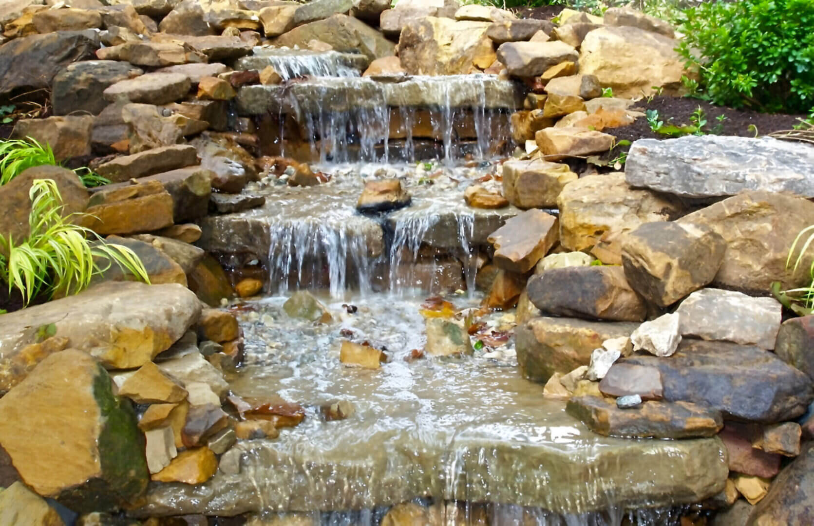 A small, multi-tiered waterfall flows over rocks, surrounded by green plants and moss.