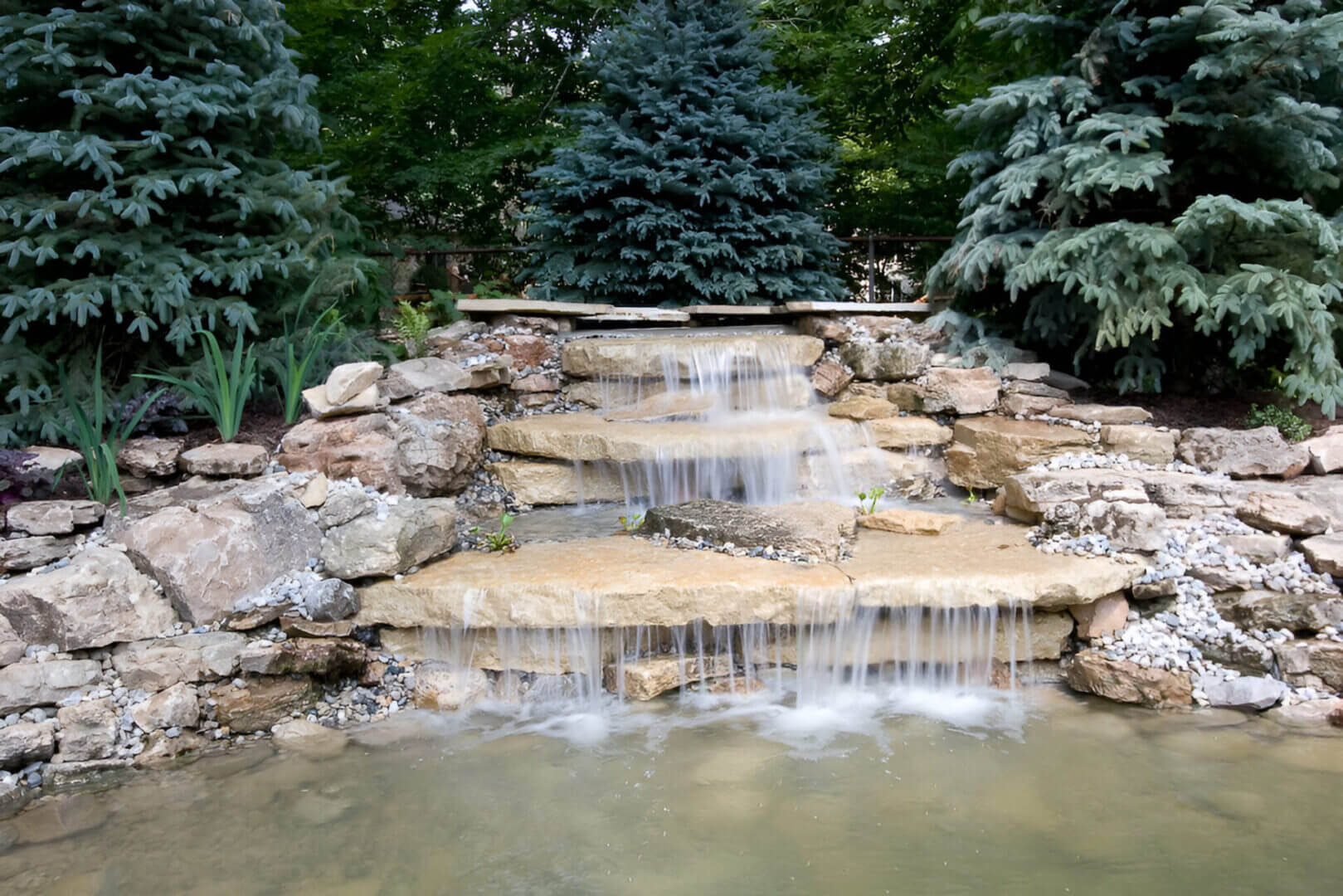 Small artificial waterfall cascading over stone steps, surrounded by rocks and green pine trees, leading into a clear pond below.