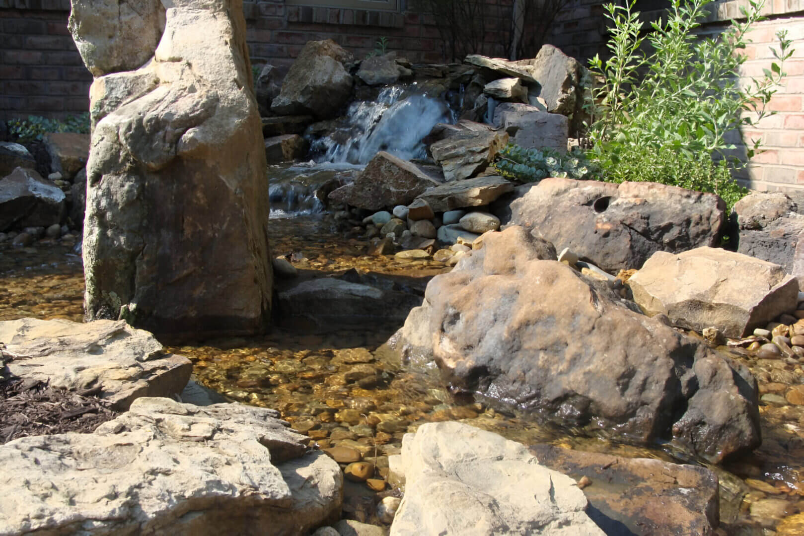 A small waterfall flows over rocks into a shallow, pebble-filled pond surrounded by greenery and stone.