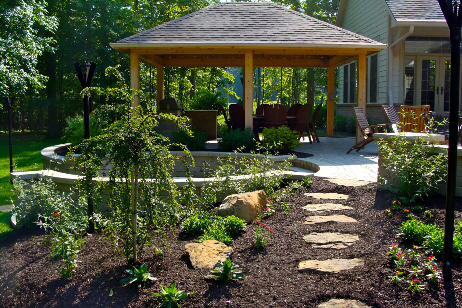 Garden path leading to a wooden gazebo surrounded by plants and trees, with patio furniture inside.
