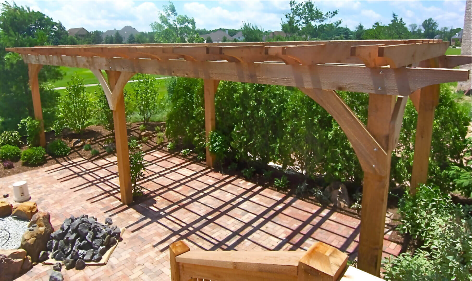 Wooden pergola on a sunny patio with brick flooring, surrounded by lush green plants and trees.